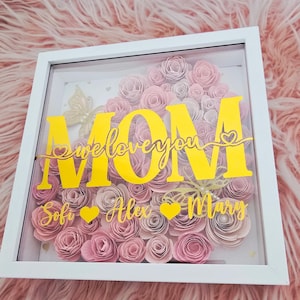 Puede incluir: Una obra de arte floral enmarcada con un borde blanco. La obra presenta una disposición en forma de corazón de rosas de papel rosa. La palabra "MOM" se muestra en amarillo, junto con el texto "we love you" y los nombres "Sofi, Alex, Mary".