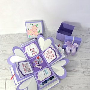 May include: A handmade, open explosion box with purple and white heart-shaped panels. The box contains decorative cards with phrases like "Love You Mom" and a keychain. Additional purple gift boxes with floral and bow accents are also present.