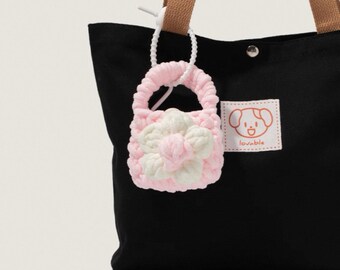 May include: A black tote bag with brown straps and a small, pink and white crocheted purse charm. The charm is shaped like a small handbag with a flower design. A white tag with a dog illustration and the word "lovable" is attached to the bag.