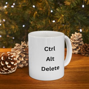 May include: White ceramic mug with the text "Ctrl Alt Delete" in black. The mug is on a wooden surface with pine cones and a blurred background of green foliage and lights.