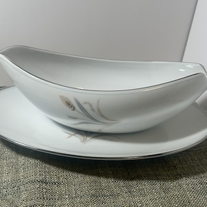 May include: White gravy boat with a matching saucer. The boat has a curved design with a silver trim. The saucer also has a silver trim and a wheat-like design in the center. The set is on a gray surface.