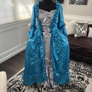 May include: A teal and silver historical costume dress on a mannequin. The dress features a ruffled bodice, long sleeves, and a full skirt. Silver bows and lace trim add detail to the elegant design. The dress is made of a shiny fabric.