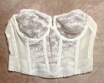 May include: A white, vintage-style bustier with delicate floral lace detailing. The bustier features structured cups and a fitted bodice, with sheer panels and a scalloped hemline. The overall design is elegant and romantic.