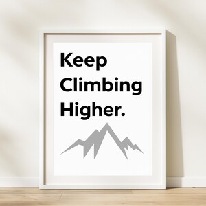 May include: White framed print with the text "Keep Climbing Higher." in black, above a gray mountain graphic. The print is displayed on a light wood surface.