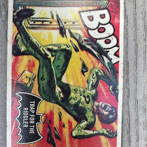 Könnte beinhalten: Vintage Sammelkarte mit einer grünen Figur in dynamischer Pose vor rotem und gelbem Hintergrund. Das Wort "BOOM" ist prominent dargestellt, mit dem Text "TRAP FOR THE RIDDLER" in einem fledermausförmigen Emblem.