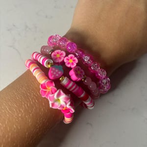 May include: A collection of pink bracelets with various designs. One bracelet features clear beads, while others have flower and strawberry charms. The bracelets are made of colorful beads and polymer clay, with a mix of solid and striped patterns.