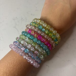 May include: A collection of colorful, beaded bracelets in shades of pink, blue, green, yellow, and clear. The bracelets are made of small, round beads and are stacked on a wrist.