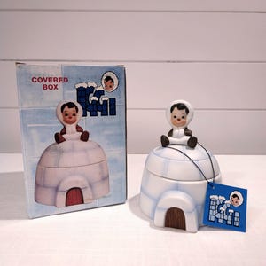 May include: A ceramic igloo-shaped covered box with a small figure on top, next to its matching box. The box and figure are white with blue accents and a brown door. The box has the words "COVERED BOX" printed on it.