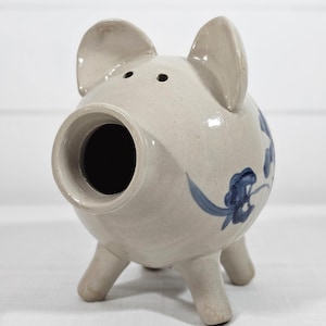 May include: A ceramic piggy bank in a light beige color with blue floral accents. The bank has a large snout, two small ears, and three legs. The coin slot is located on the top of the head.