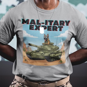 May include: A light gray t-shirt featuring a cartoon illustration of a German Shepherd dog wearing goggles and a military uniform, riding a green tank in a desert landscape. The text "MAL-ITARY EXPERT" is printed above the image.