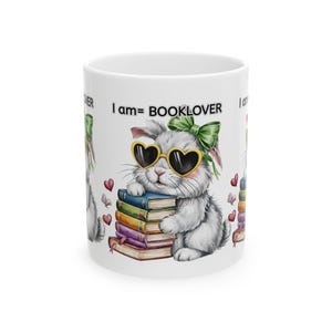 May include: White ceramic mug with a cartoon rabbit wearing heart-shaped sunglasses and a green bow. The rabbit is holding a stack of books, with the text "I am= BOOKLOVER" above it. Red hearts and butterflies are scattered around the design.