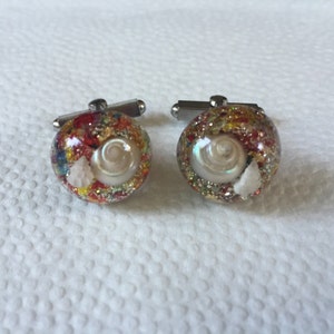 May include: A pair of cufflinks with a round, clear resin design. Each cufflink features a small seashell and colorful glitter embedded in the resin.