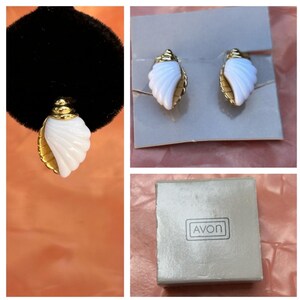 May include: A pair of gold-tone earrings shaped like white seashells with ribbed textures, presented on a gray display card alongside a square box featuring the brand name AVON printed in black text on the top surface of the packaging.