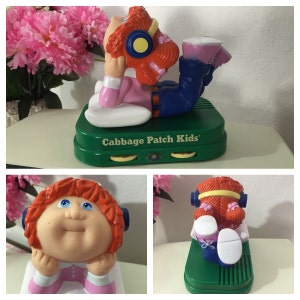 May include: A Cabbage Patch Kids toy with red hair wearing headphones and a pink shirt. The toy is sitting on a green base with the words "Cabbage Patch Kids" printed on it.