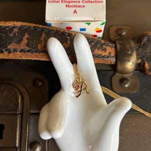 May include: A gold-colored initial necklace with a red gem, displayed on a white ceramic hand. The necklace is from the Avon Kids Initial Elegance Collection. The box is white with colorful accents.