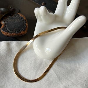 May include: A gold-colored herringbone chain necklace. The necklace is displayed on a white ceramic hand-shaped jewelry holder. The background includes a vintage trunk with a leather clasp.