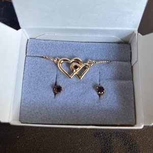 May include: A gold necklace and earrings set featuring two interlocking hearts with a red gemstone in the center of each heart. The jewelry is displayed in a white box.