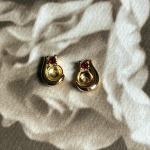 May include: A pair of gold-toned stud earrings, each featuring a smooth, teardrop-shaped metal frame with a circular cutout in the center and a single, small, round, deep red gemstone set at the top point of the earring design.