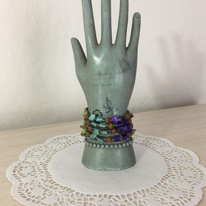 May include: A stack of colorful beaded bracelets on a hand-shaped display stand. The bracelets are made of turquoise, brown, and purple beads.