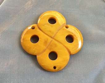 May include: A yellow, four-leaf clover shaped pendant with four holes. The pendant is made of plastic and has a smooth, glossy finish.