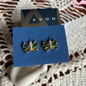 May include: A pair of butterfly earrings with green and iridescent accents, displayed on a blue card. The earrings are set against a white ceramic hand-shaped holder, with an Avon box in the background.