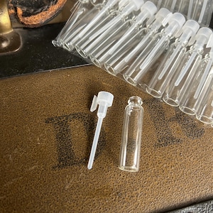 May include: Clear glass perfume sample vials with white plastic caps and applicators. A collection of vials is packaged in a clear plastic bag. One vial and applicator are separate. The vials are on a brown textured surface.
