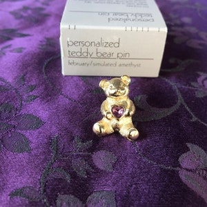 May include: Gold teddy bear pin with a purple simulated amethyst heart. The pin is packaged in a white box with the text "personalized teddy bear pin february/simulated amethyst".
