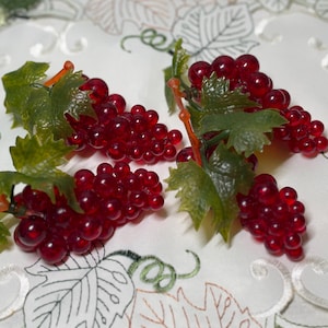 May include: Three clusters of artificial red grapes made from a translucent, glossy material. Each bunch features small, round, deep red berries attached to a short stem with several textured, green plastic leaves extending outward.