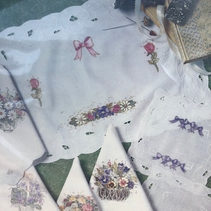 May include: Set of white embroidered napkins with floral and bow designs. The napkins feature a variety of flowers, including roses, pansies, and daisies. The napkins are perfect for adding a touch of elegance to any table setting.