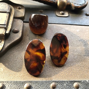 May include: A set of three tortoise shell-patterned resin jewelry pieces: a ring and two earrings. The ring is wide and the earrings are oval shaped.