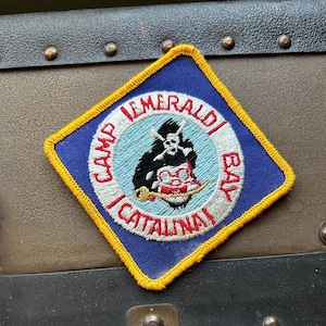 May include: A blue and yellow embroidered patch with a white circle containing the text "Camp Emeraldi Bay I Catallina" and a black and white illustration of a pirate with a skull and crossbones.