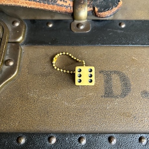 May include: A small, yellow die charm with black dots hangs from a gold-colored chain. The die is positioned on a vintage trunk with a leather handle and metal hardware. The trunk has a brown and black color scheme.