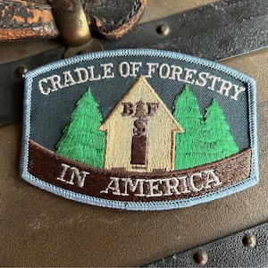 May include: A blue and brown embroidered patch with the text "CRADLE OF FORESTRY IN AMERICA" and a small cabin with the text "BIF" above the door. The cabin is surrounded by two green trees.