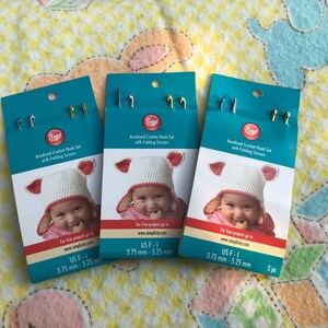 May include: Three packages of anodized crochet hooks with folding scissors. Each package features a baby wearing a crocheted hat with ear flaps and the text "Beye Anodized Crochet Hook Set with Folding Scissors". The package also includes the size of the hooks: "US F-1 3.75 mm - 5.25 mm".