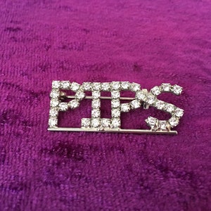 May include: A silver-toned brooch with the word "PIPS" spelled out in rhinestones. The brooch is on a purple velvet background.