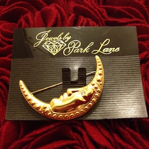 May include: A gold-toned crescent moon brooch with a sleeping face and a star, set with a small clear gem. The brooch is on a black card with the text "Jewels by Park Lane". The background is a textured red fabric.