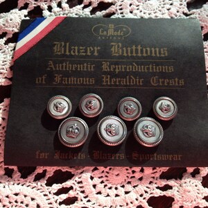May include: A set of eight silver blazer buttons with a white enamel finish and a crown design. The buttons are packaged on a black card with the text "La Mode Buttons Authentic Reproductions of Famous Heraldic Crests for Jackets Blazers-Sportswear".