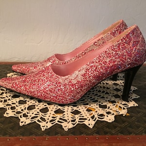 May include: A pair of pink high-heeled shoes with a sparkly, lace-like design. The shoes have a pointed toe and a thin heel.
