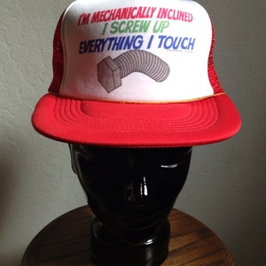 May include: Red and white trucker hat with a mesh back. The hat has a white front panel with the text "I'm mechanically inclined I screw up everything I touch" in colorful letters. A gray screw is pictured on the hat.