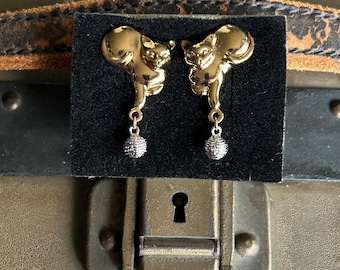 May include: A pair of gold-colored cat earrings. Each earring features a cat-shaped charm with a dangling silver-colored ball. The earrings are displayed on a black velvet card.