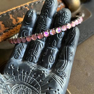 May include: A silver bracelet with pink iridescent rhinestones. The bracelet is on a black hand with various designs.