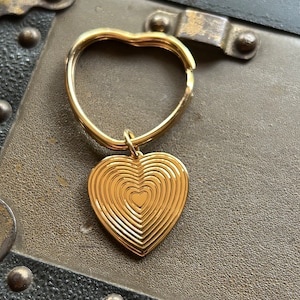 May include: A gold-colored heart-shaped keychain with a concentric circle design. The keychain is attached to a gold-colored ring.