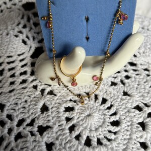 May include: A set of three gold-toned jewelry pieces with pink gemstones. The set includes a hoop earring, a nose ring, and a delicate chain necklace.