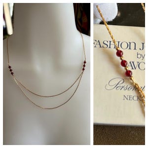 May include: A gold chain necklace with two strands and small red beads. The necklace is displayed on a white background and comes with a box that says "Fashion Jewelry by Avon Personal Necklace".