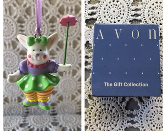 Pode incluir: Uma figurinha de coelho branco vestindo um vestido verde e roxo com bolinhas brancas, segurando uma flor rosa em um caule verde. A figurinha é um ornamento com uma fita roxa. O ornamento está embalado em uma caixa azul com o texto "AVON The Gift Collection".