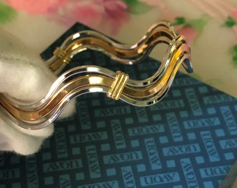 May include: A wavy, two-tone bracelet featuring alternating silver and gold-colored bands with small, vertical gold-colored accents. The bracelet is positioned above a blue box with a repeating pattern of the word AVON printed in white.