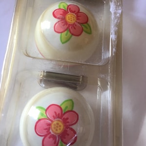 May include: Two white ceramic cabinet knobs with a pink flower design. The flower has a yellow center and green leaves.