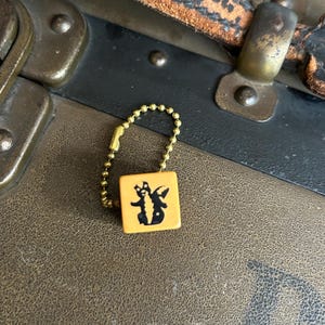 May include: A small, square, orange charm with a black graphic, attached to a gold-colored ball chain. The charm is set against a backdrop of a vintage brown leather trunk with brass hardware.