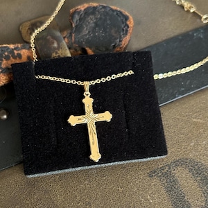 May include: A gold-colored cross pendant necklace displayed on a black velvet surface. The cross has an ornate design with etched details and is suspended from a delicate chain. The necklace is presented on a textured, brown surface.