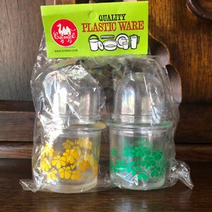 May include: Two clear glass spice jars with silver lids, each wrapped in plastic. One jar has yellow flower designs, the other has green flower designs. A green and yellow label reads "Quality Plastic Ware" with a camel logo.